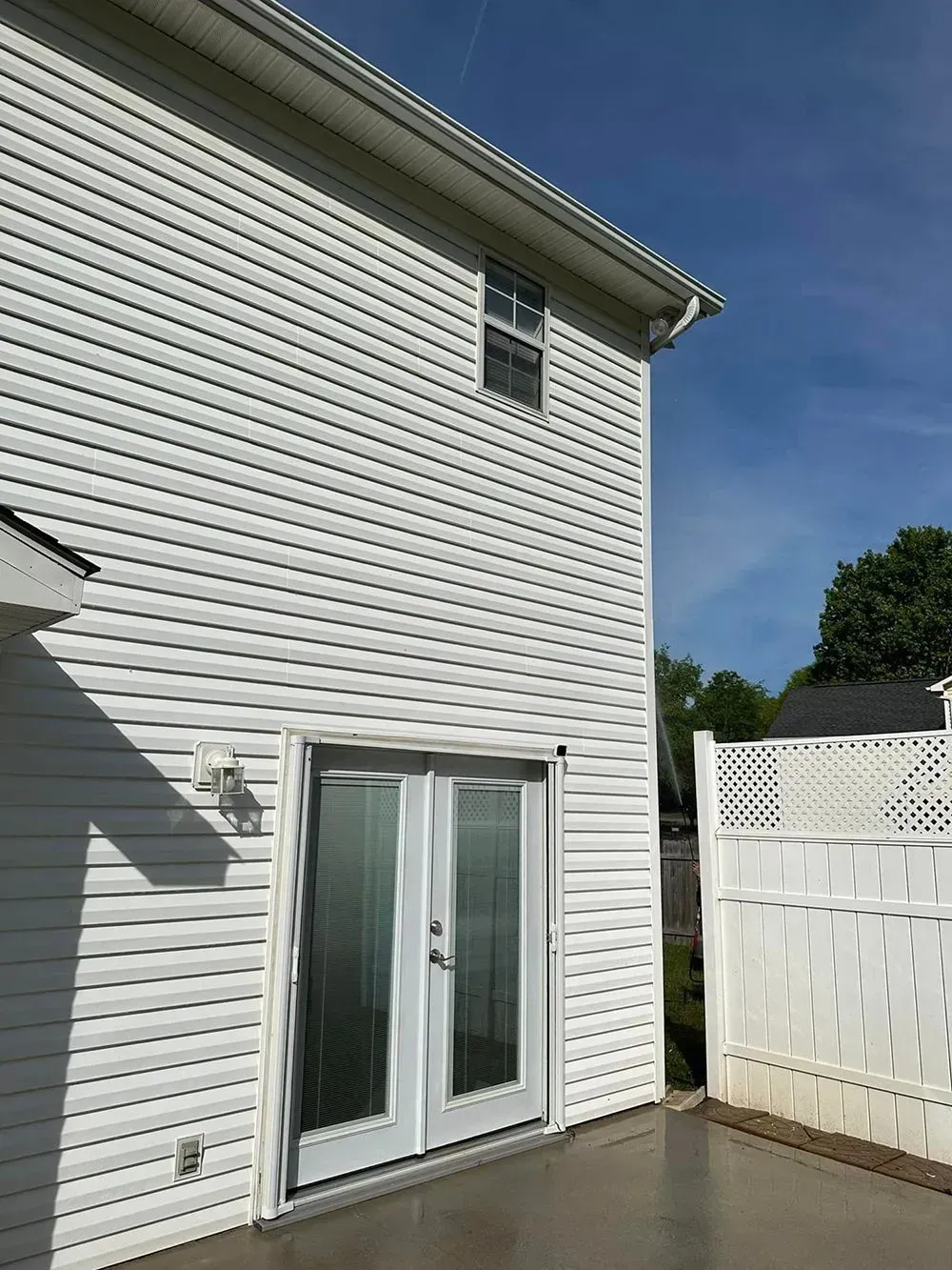 A view of a two-story house exterior with white siding, French doors, and a white fence under a blue sky.