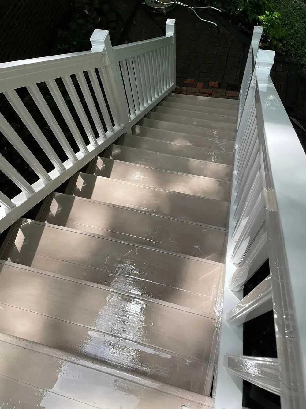 A high-angle view of wet, light-brown outdoor stairs with white railings leading down into a shaded area.
