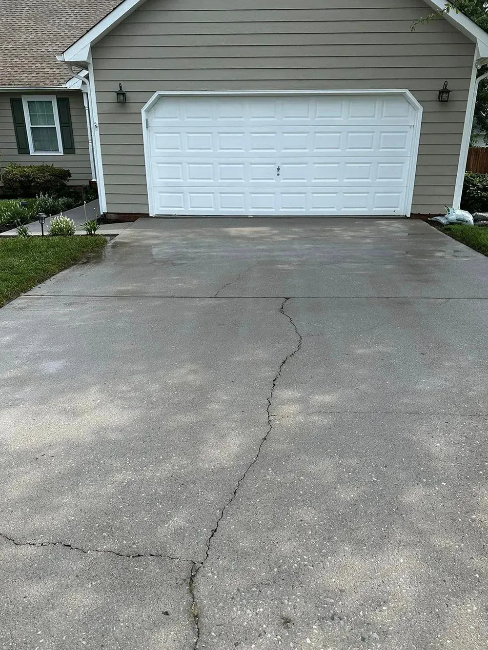 A beige house with a white garage door features a driveway with a prominent crack running through the concrete.