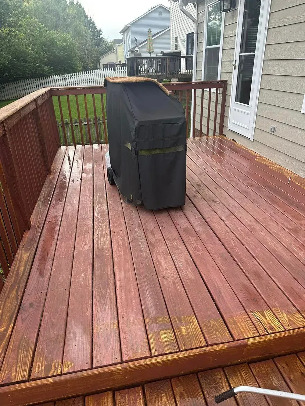A covered grill sits in the middle of a weathered, reddish-brown wooden deck outside a house.