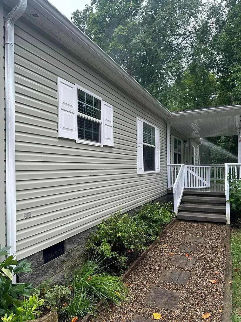 Tan vinyl-sided house with white shutters and trim, featuring a wooden deck with stairs and a pea gravel walkway.