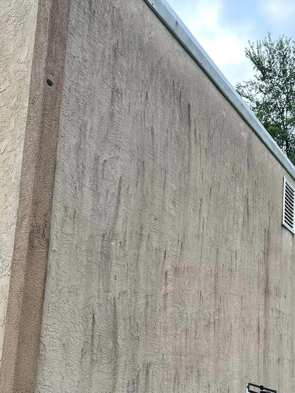 An exterior wall with a textured, beige stucco finish featuring vertical dark streaks from water runoff.