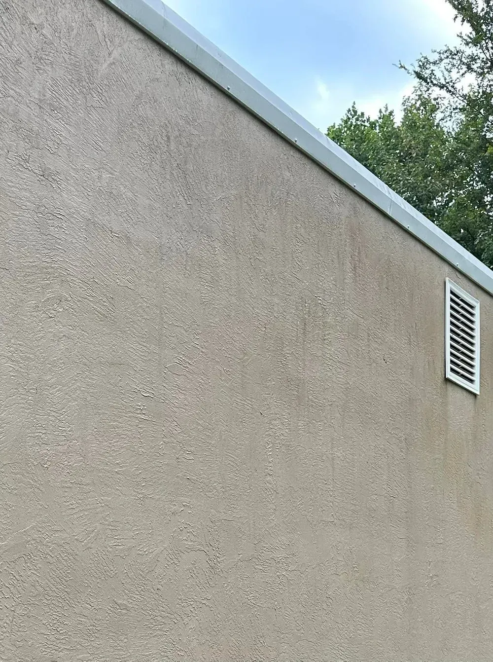 A tan textured exterior wall with a horizontal white gutter at the top and a small rectangular ventilation grate.