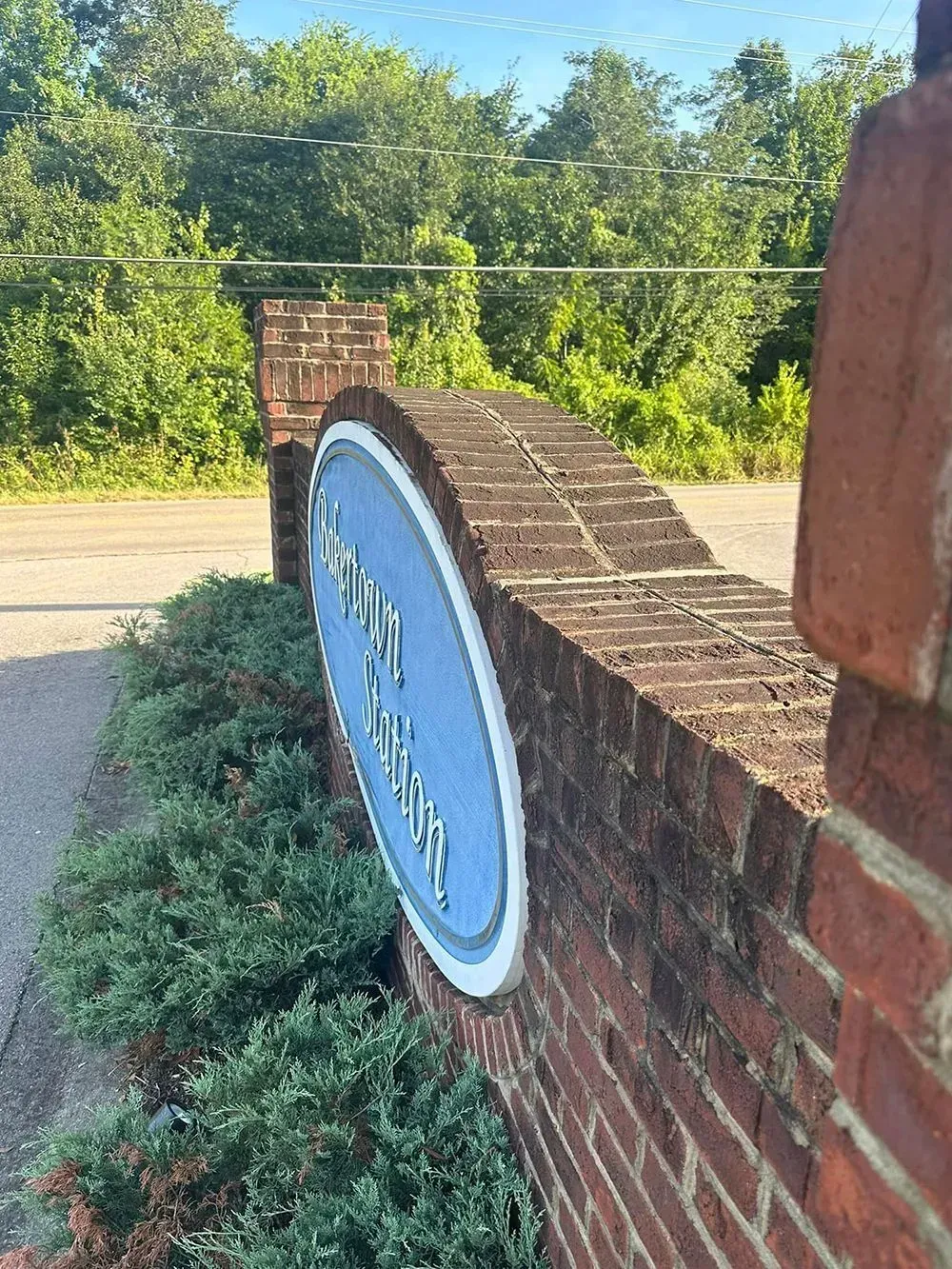 A brick wall with a blue oval sign partially visible, set against a background of lush green trees and a paved road.