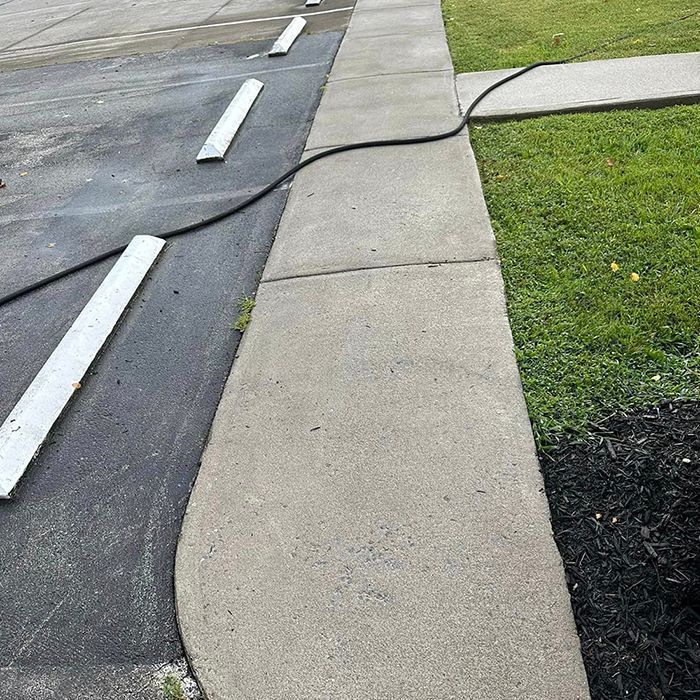 A long black power cord lies across a concrete sidewalk and asphalt parking area adjacent to a grassy lawn.