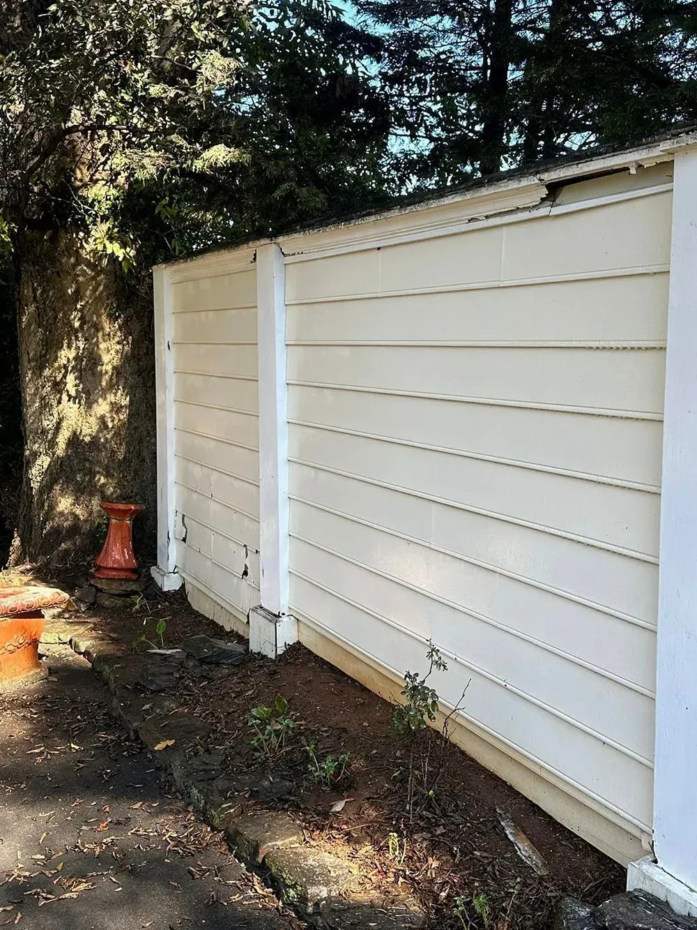 A beige, horizontal-paneled wall with white vertical posts stands beside a dirt patch with small plants and a red object.