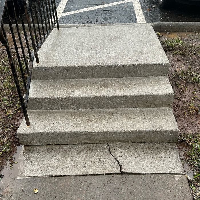 A set of three concrete steps leading to an elevated landing, with a black metal handrail on the left.