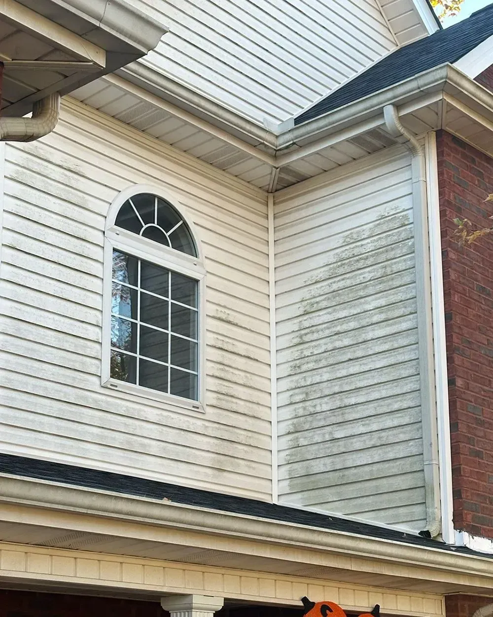 A corner of a house exterior showing light-colored vinyl siding with significant dark, streaky grime and algae buildup.
