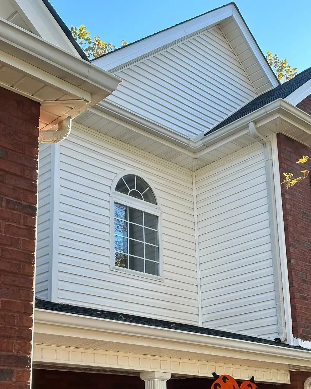 A cream-colored vinyl-sided upper story of a house with an arched window, set between two brick walls under a gabled roof.