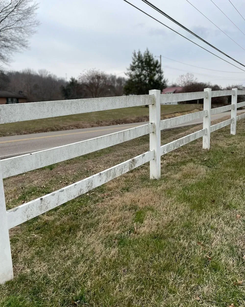 A white, three-rail wooden fence borders a rural road under an overcast sky.