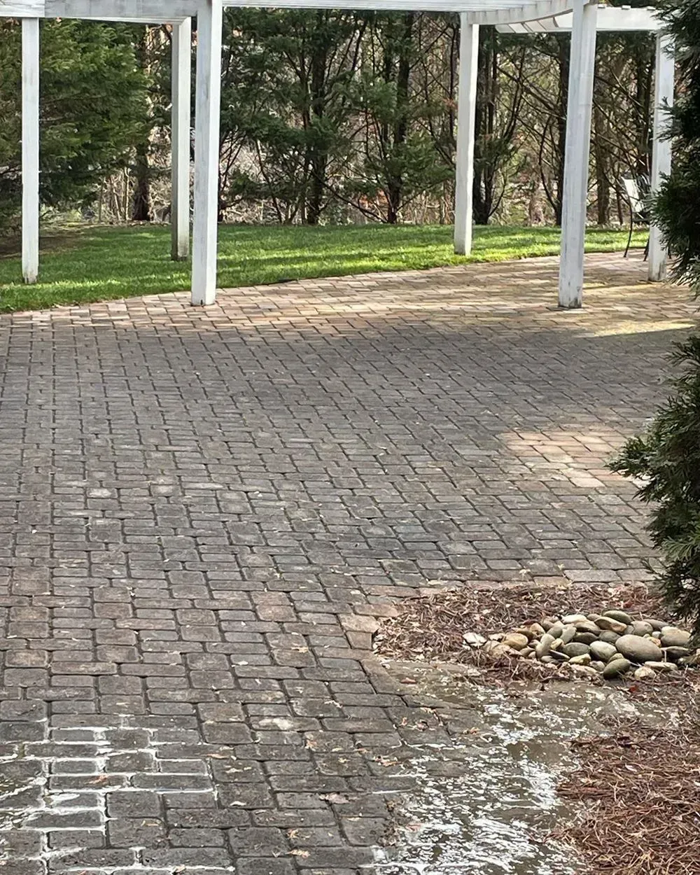 A brick patio under a white pergola, bordered by green grass and trees with a small pile of decorative rocks in the corner.