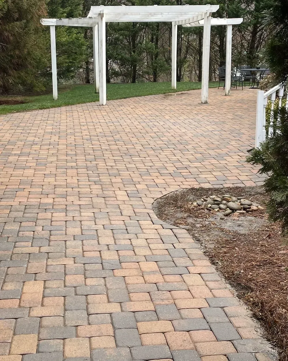 A paved patio with multi-toned rectangular stones, leading to a white pergola set on a grassy lawn.