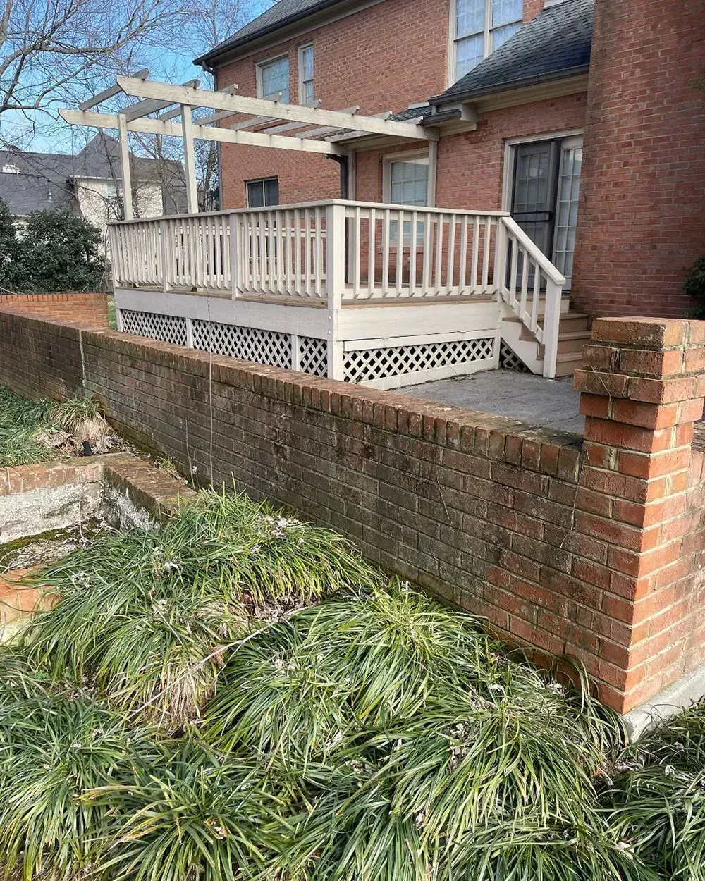 A wooden deck with a pergola and white railing sits behind a weathered red brick retaining wall in a yard.