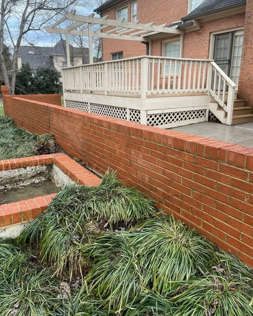 A light-colored wooden deck with a pergola sits behind a red brick garden wall in a yard with overgrown grass.