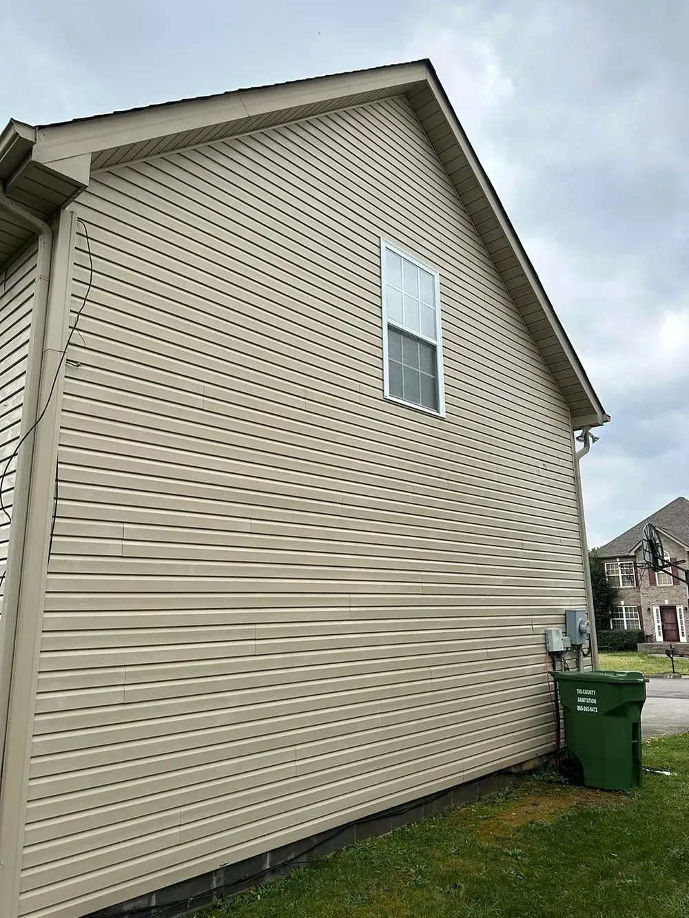 A side view of a beige house exterior with distorted, wavy vinyl siding, a single window, and a green trash bin.