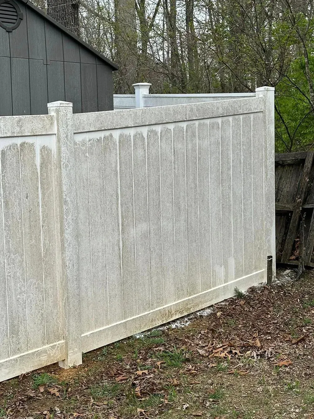 A dirty, weathered white vinyl privacy fence stands outdoors with a dark shed visible behind it.