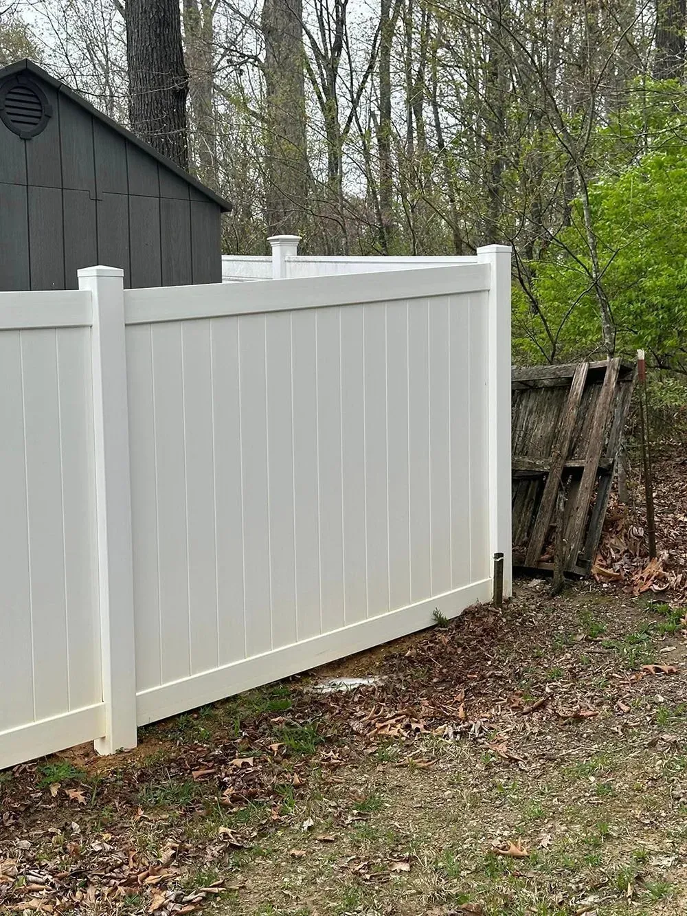 A white vinyl privacy fence stands in a wooded yard near a shed and a wooden pallet.