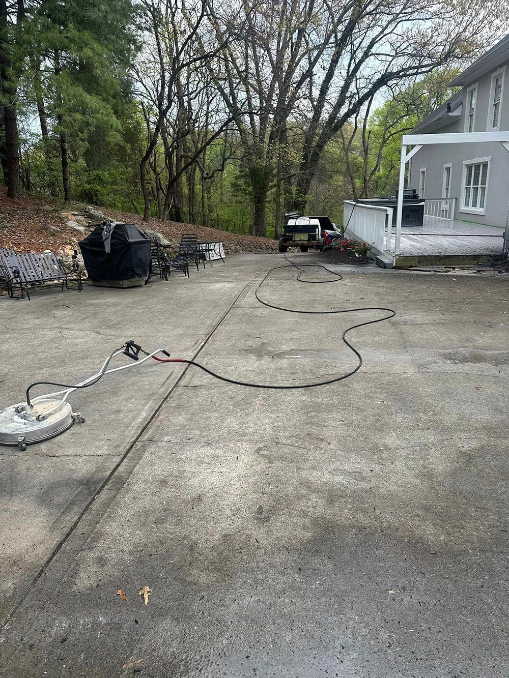 A surface cleaner with a long black hose lies on a stained concrete driveway in front of a gray house.