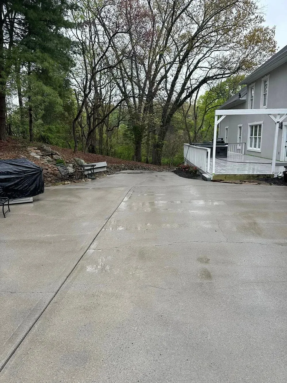 A concrete driveway leads toward a wooded area beside a house with a white deck.