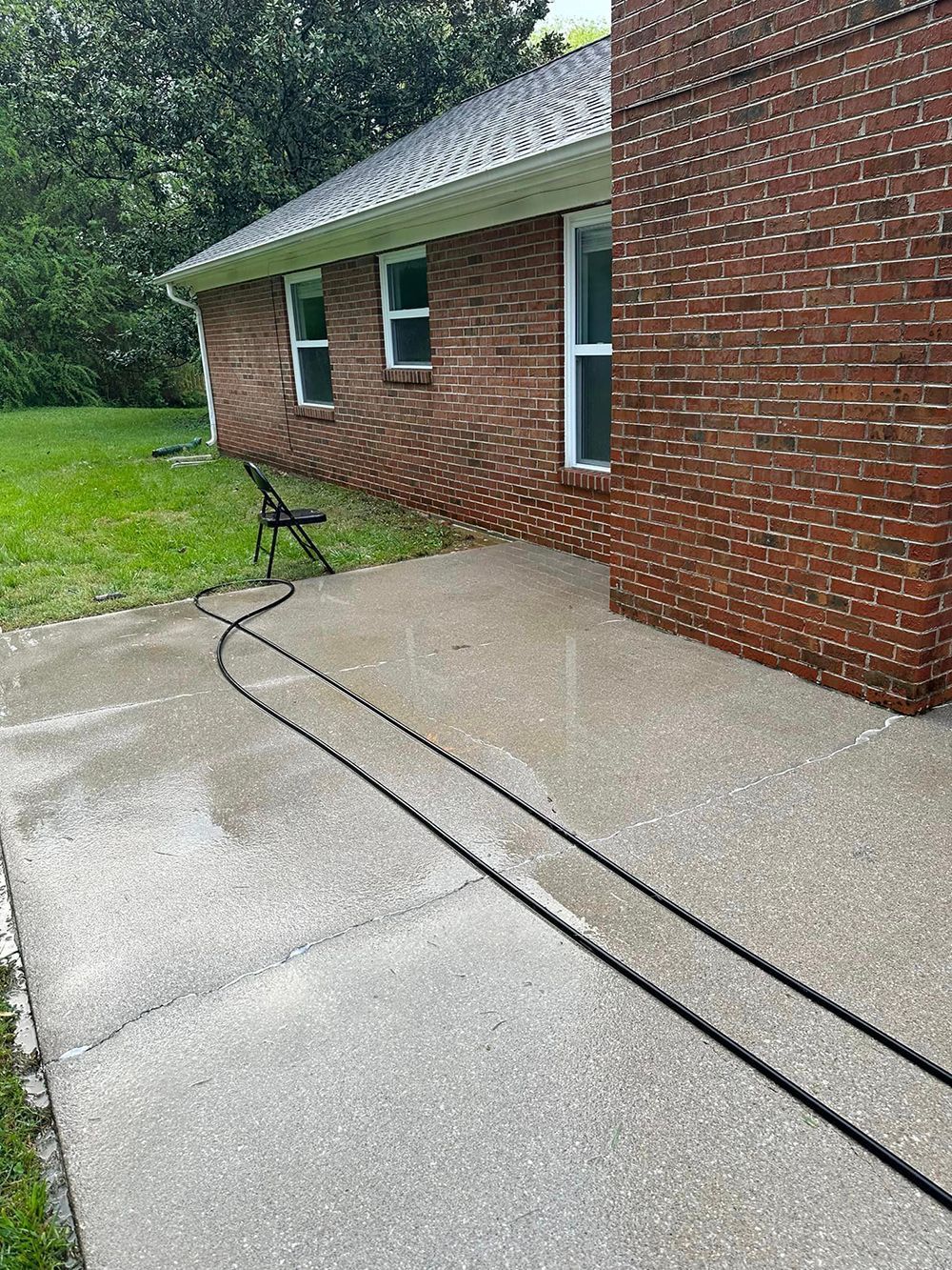 A brick house exterior with two windows and a black garden hose laying across a concrete patio in a grassy yard.