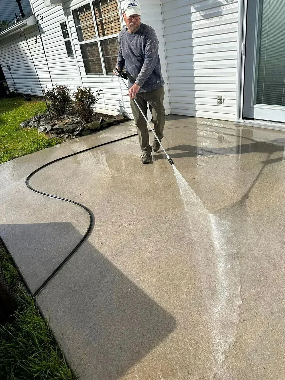 A person uses a pressure washer to clean a concrete patio next to a house with white siding.