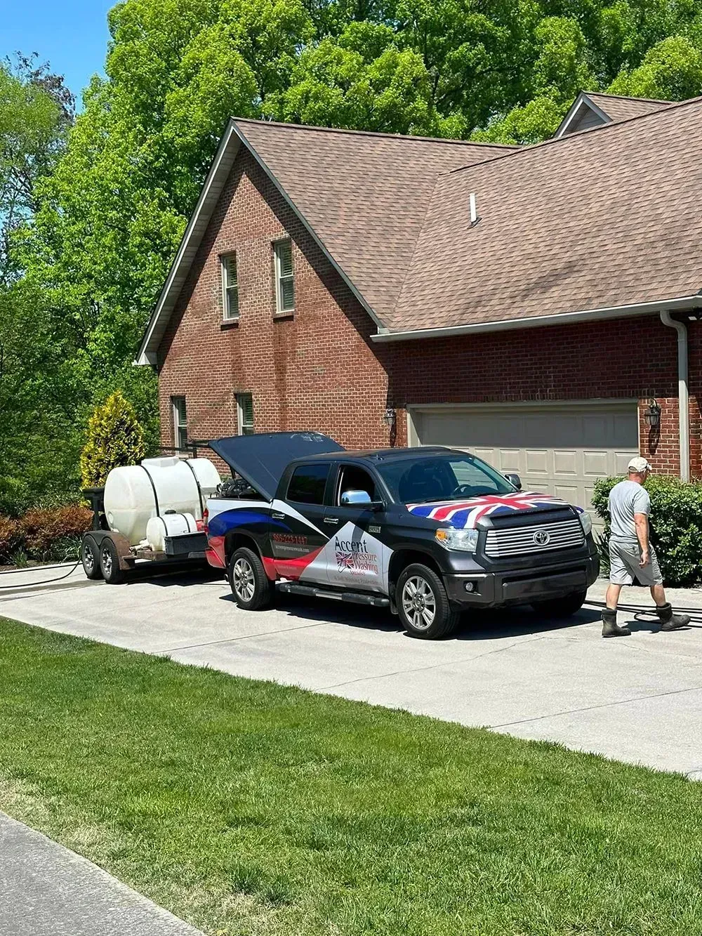 A work truck with a Union Jack hood decal and a water tank trailer parked in a residential driveway near a brick house.