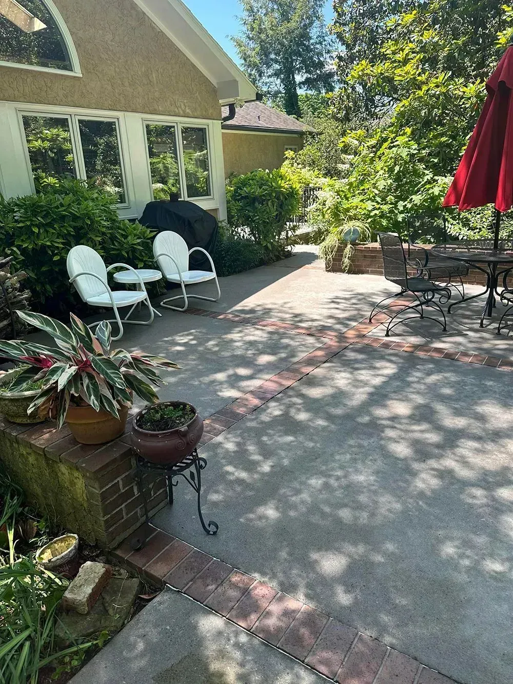 A paved patio with red brick borders, outdoor seating, potted plants, and a red umbrella next to a tan house.