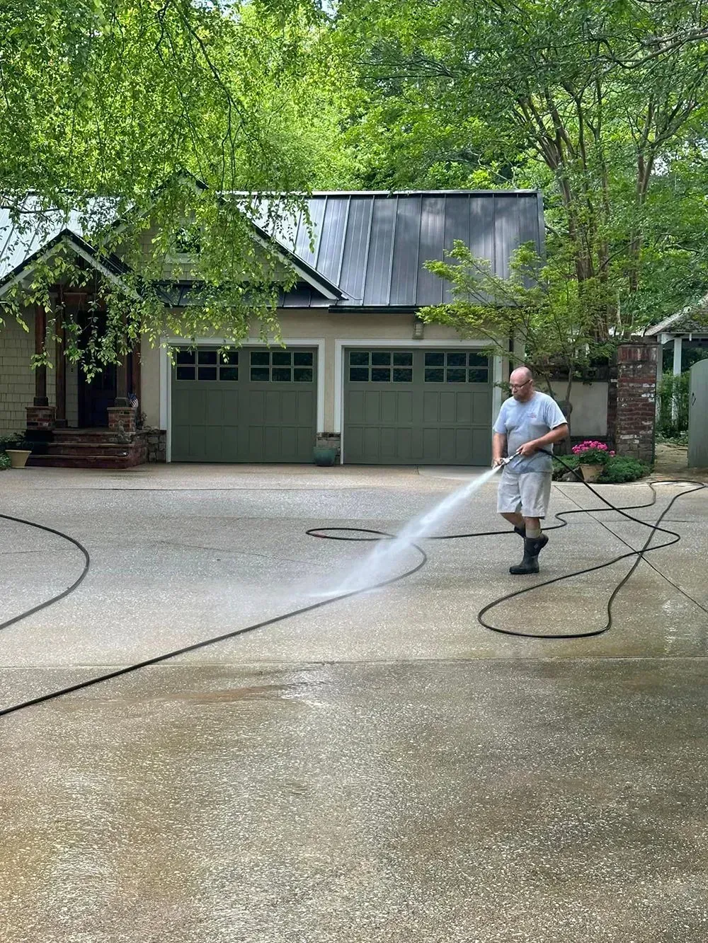 A person pressure washing a residential concrete driveway in front of a house with a green garage.