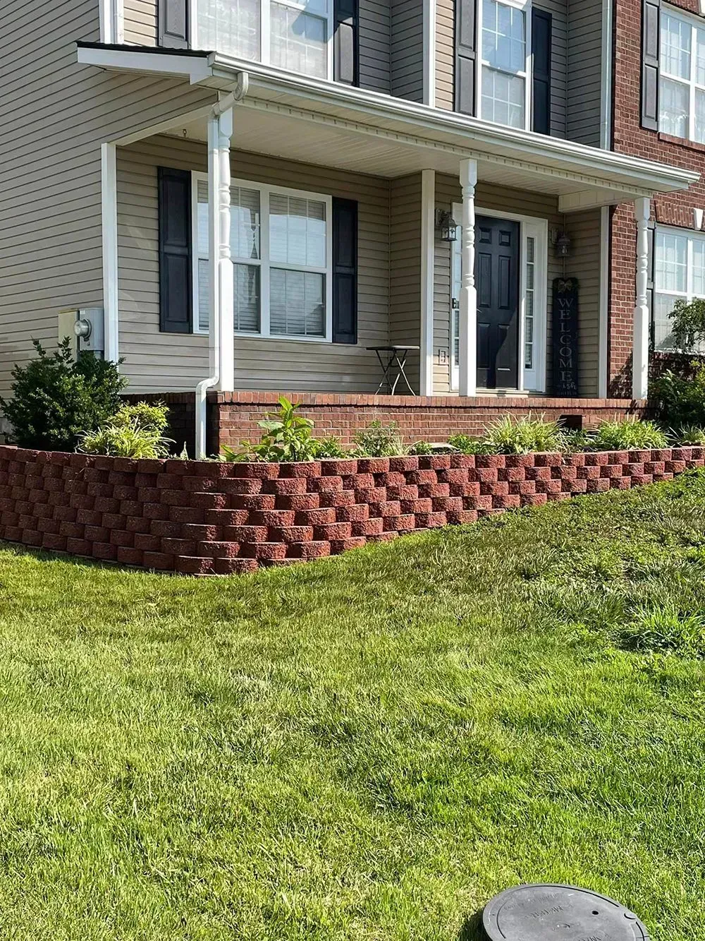 A two-story suburban house with a tan facade, black shutters, and a front porch elevated by a red brick retaining wall.