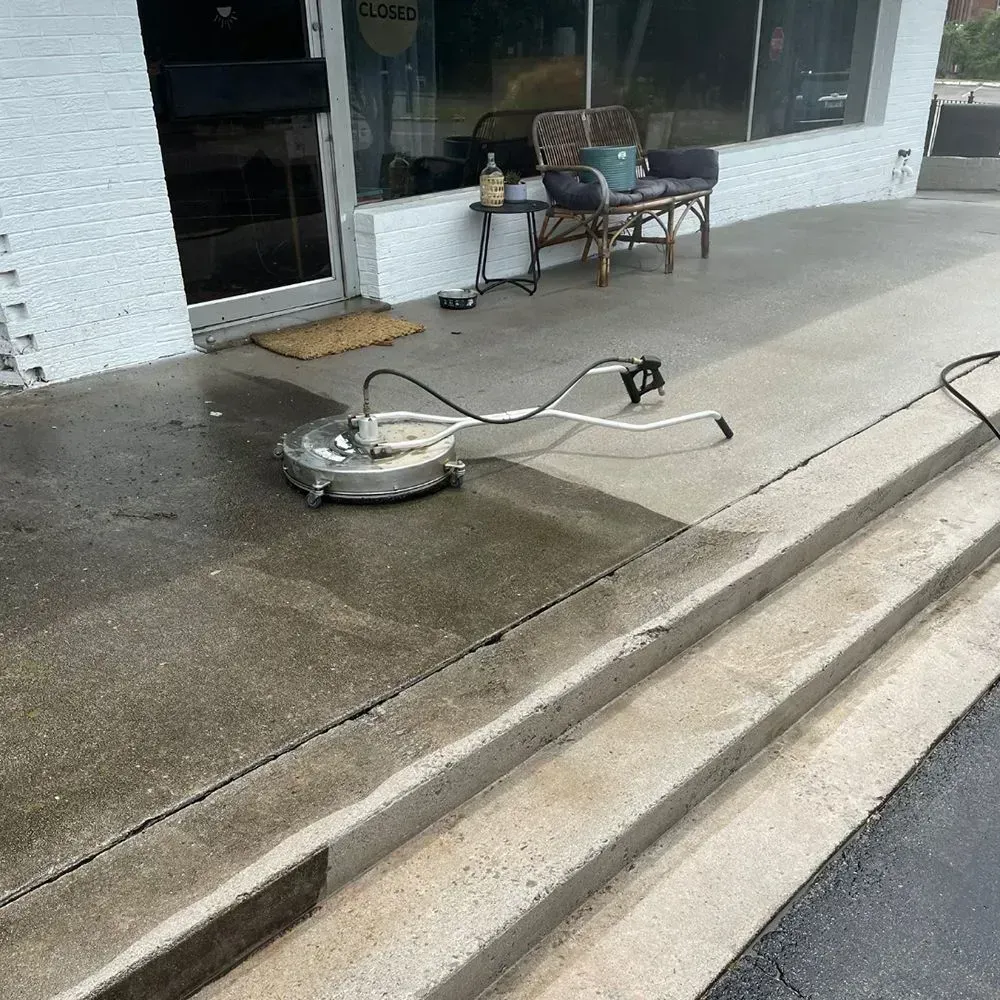 A pressure washer surface cleaner sits on a damp concrete sidewalk in front of a building entrance with outdoor furniture.