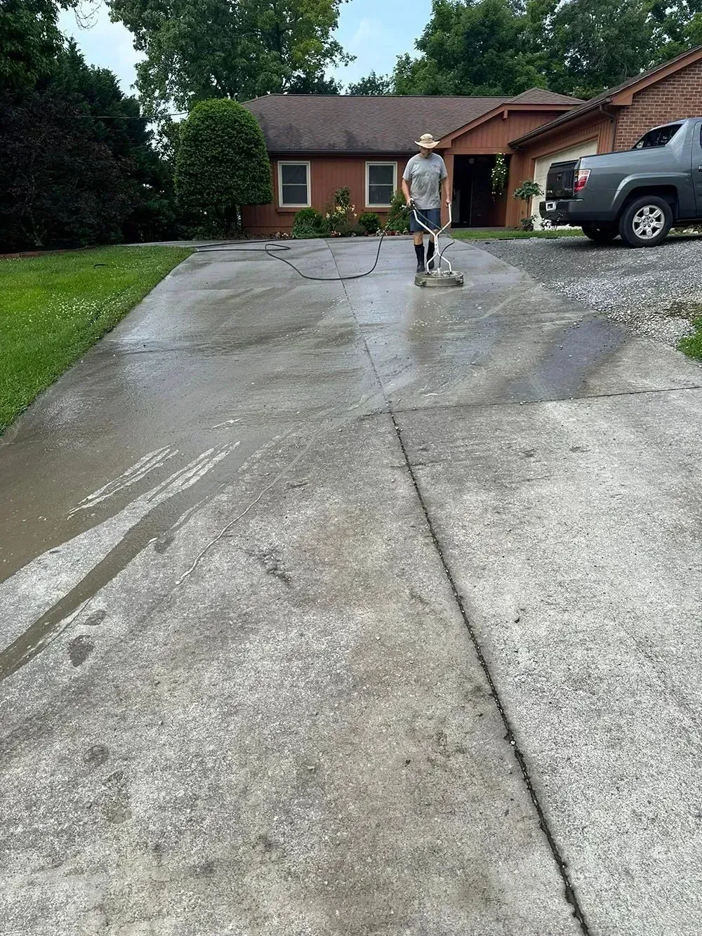 A person using a surface cleaner on a driveway in front of a house, showing a contrast between clean and dirty concrete.