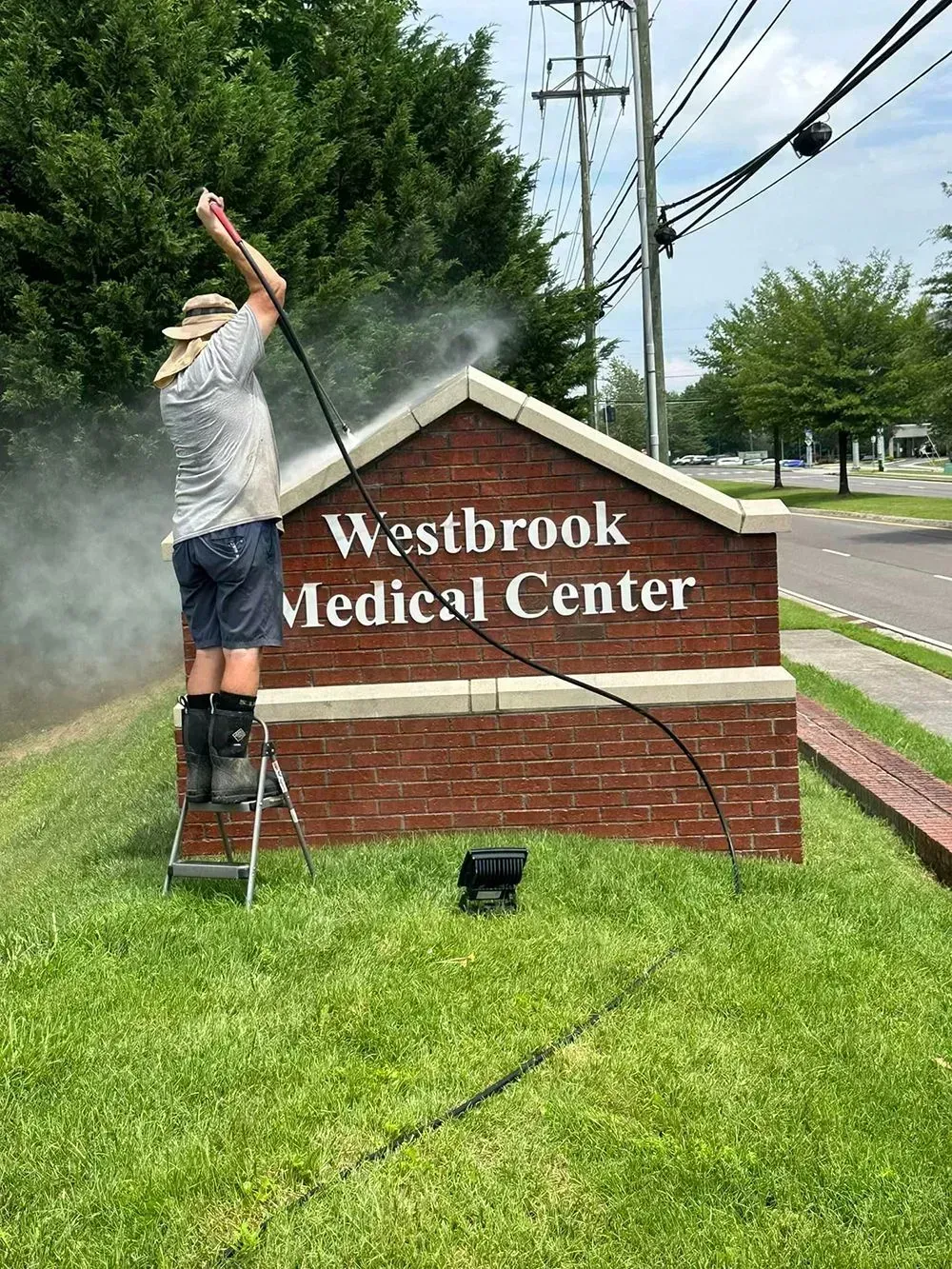 A person on a step ladder pressure washes a brick sign that reads 