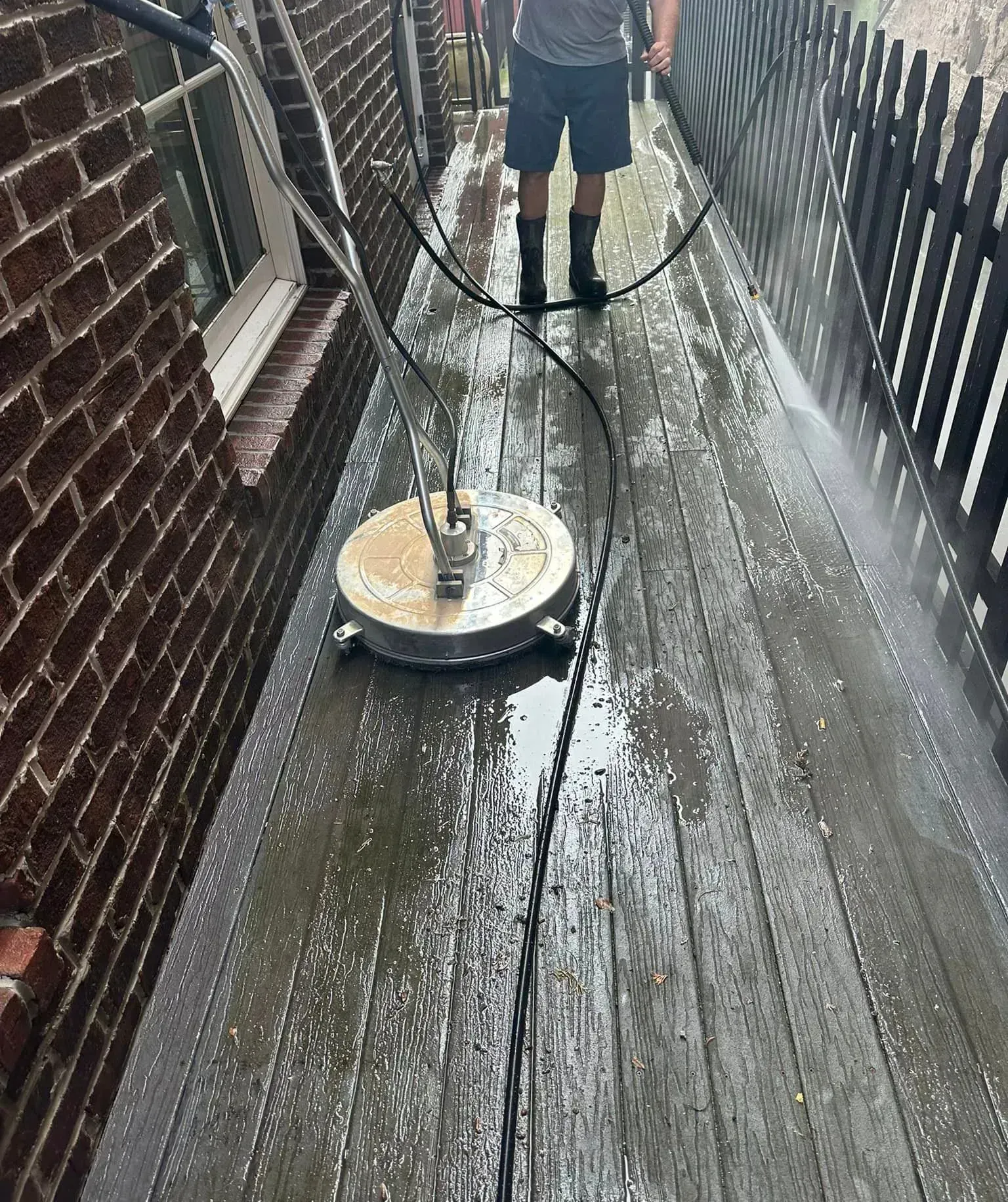 A person uses a circular power washer to clean a weathered wooden deck alongside a brick wall and black metal fence.