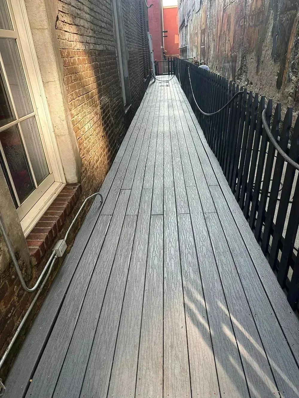 A narrow walkway with gray wooden decking leading toward a distant orange building between a brick wall and a fence.