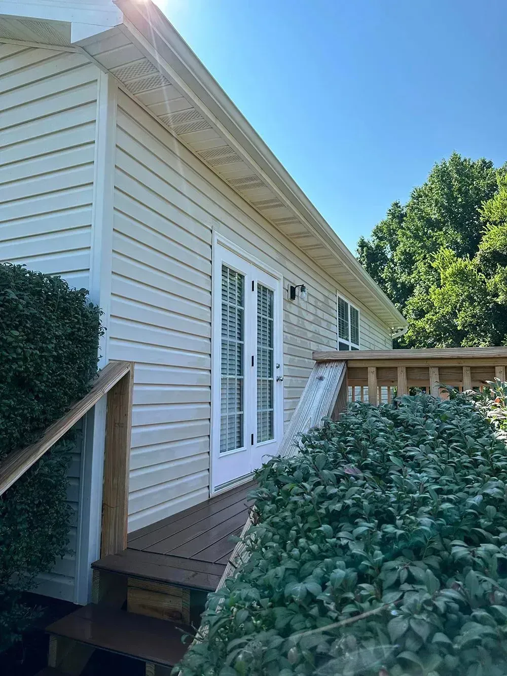 Beige siding house exterior with French doors opening onto a wooden deck with steps and lush green bushes on a sunny day.