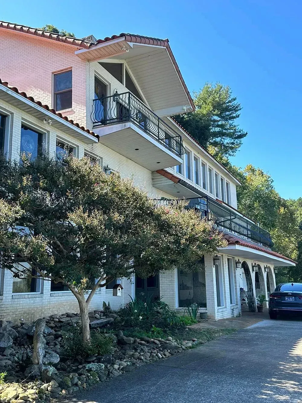 A two-story building with pink siding, a balcony, and a covered lower-level porch, featuring a tree in the foreground.