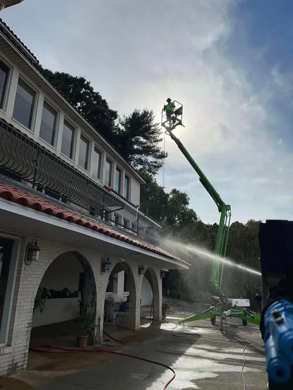 A worker in a bucket lift sprays water onto the side of a multi-story white house with arched porch columns.