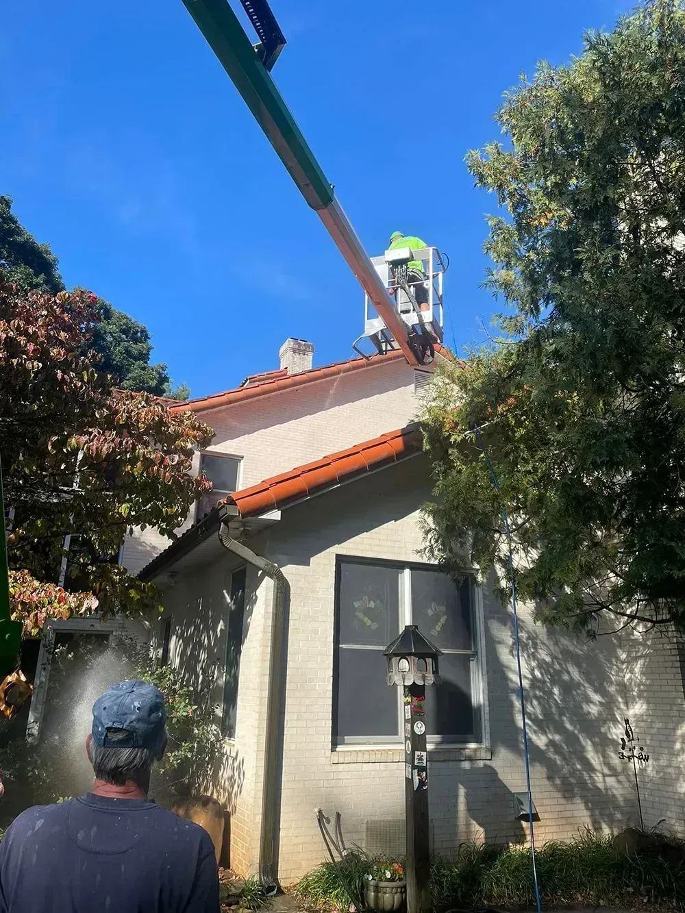 A person operates a cherry picker near a roof while another person stands in the foreground observing the work.