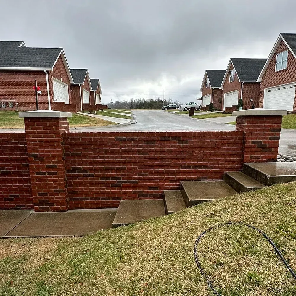 A red brick wall with two concrete-topped pillars stands before a street lined with red brick suburban houses.