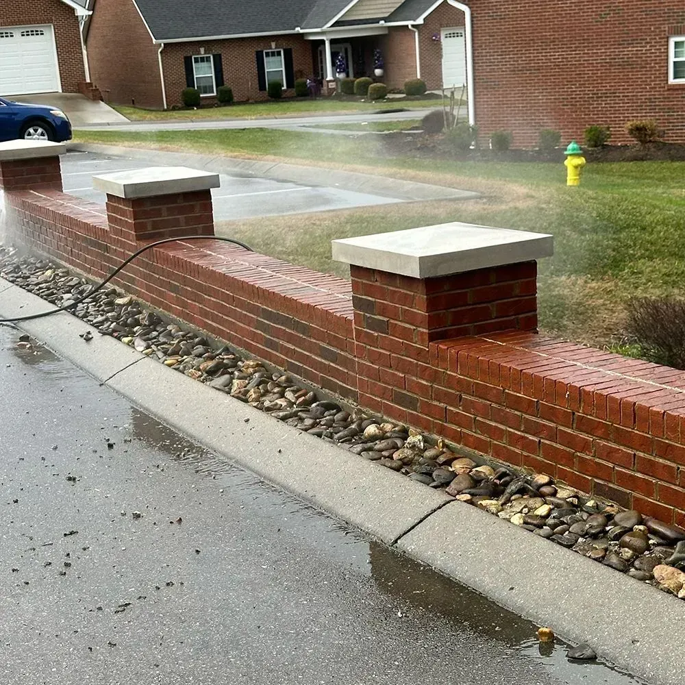 A low red brick wall with concrete pillars runs alongside a curb and gravel border in front of a residential home.