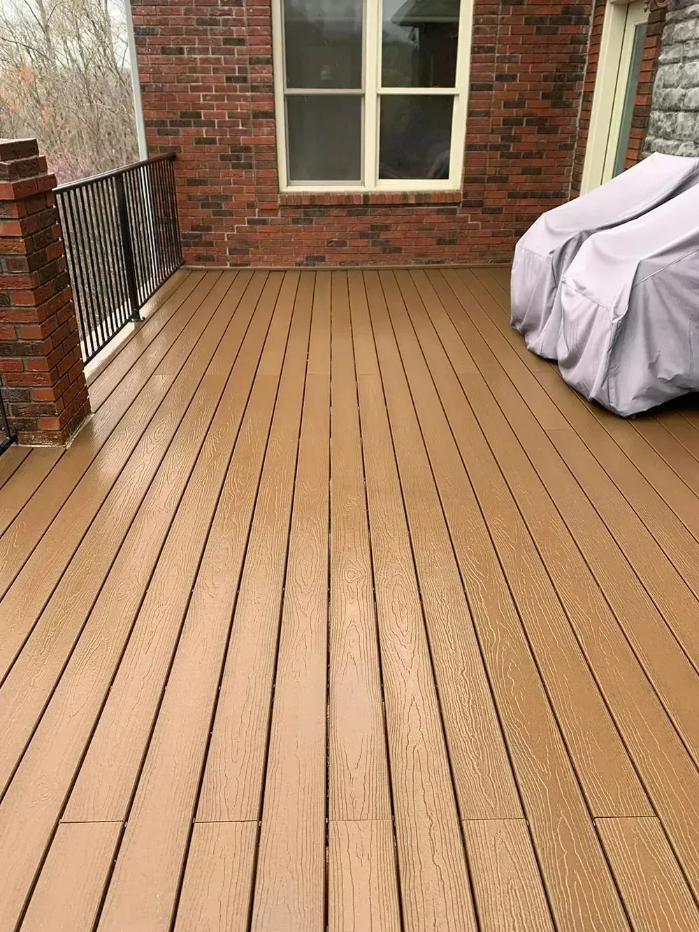 A tan, composite-plank deck attached to a brick home with an iron railing and covered furniture.