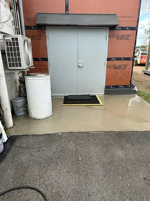 Gray double doors with an awning, black floor mat, and white barrel on a concrete pad against an unfinished exterior wall.