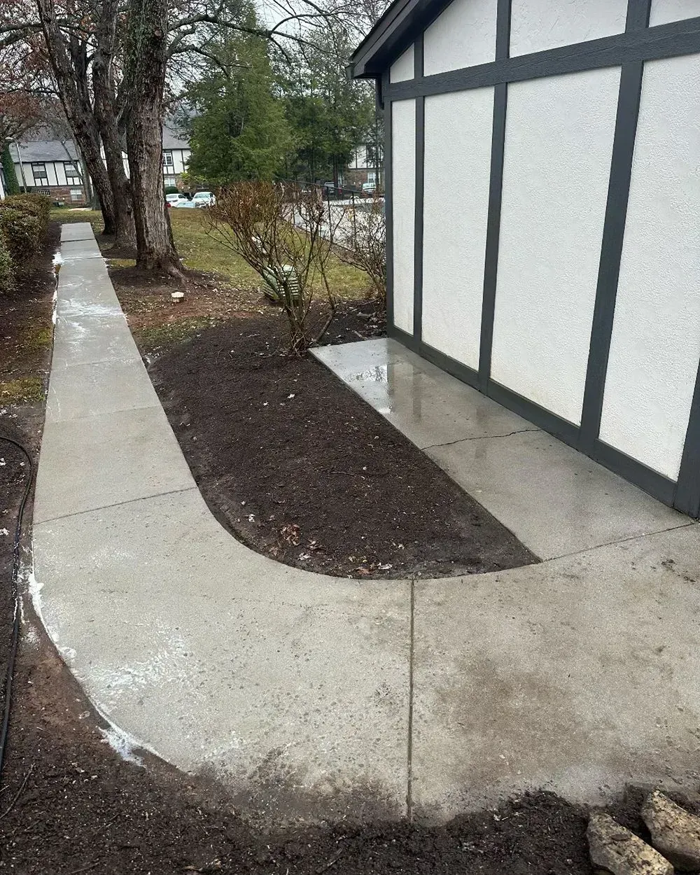 A concrete walkway curves around a mulched flower bed next to a white house with dark gray trim.