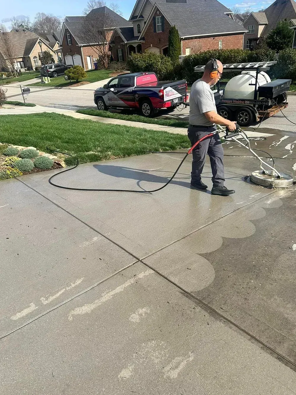 A person uses a circular surface cleaner to power wash a driveway in a residential neighborhood.