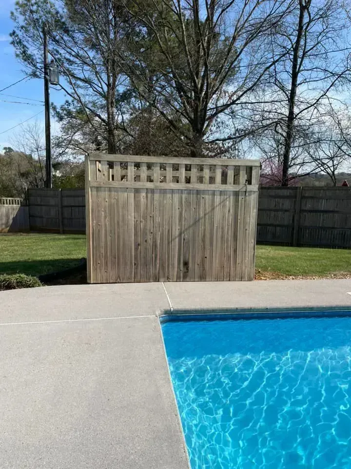 A tall wooden privacy fence stands on a lawn next to a blue swimming pool under a bright sky.