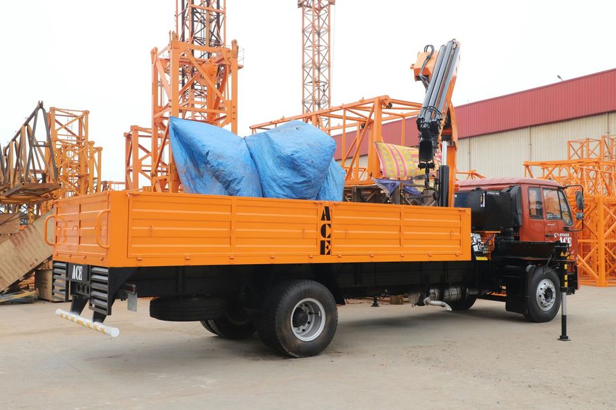 Orange flatbed truck with crane, loaded with tarp-covered materials, parked near construction equipment.