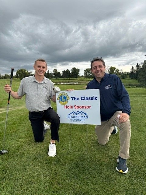 Two men are kneeling on a golf course holding a sign that says the classic hole sponsor.