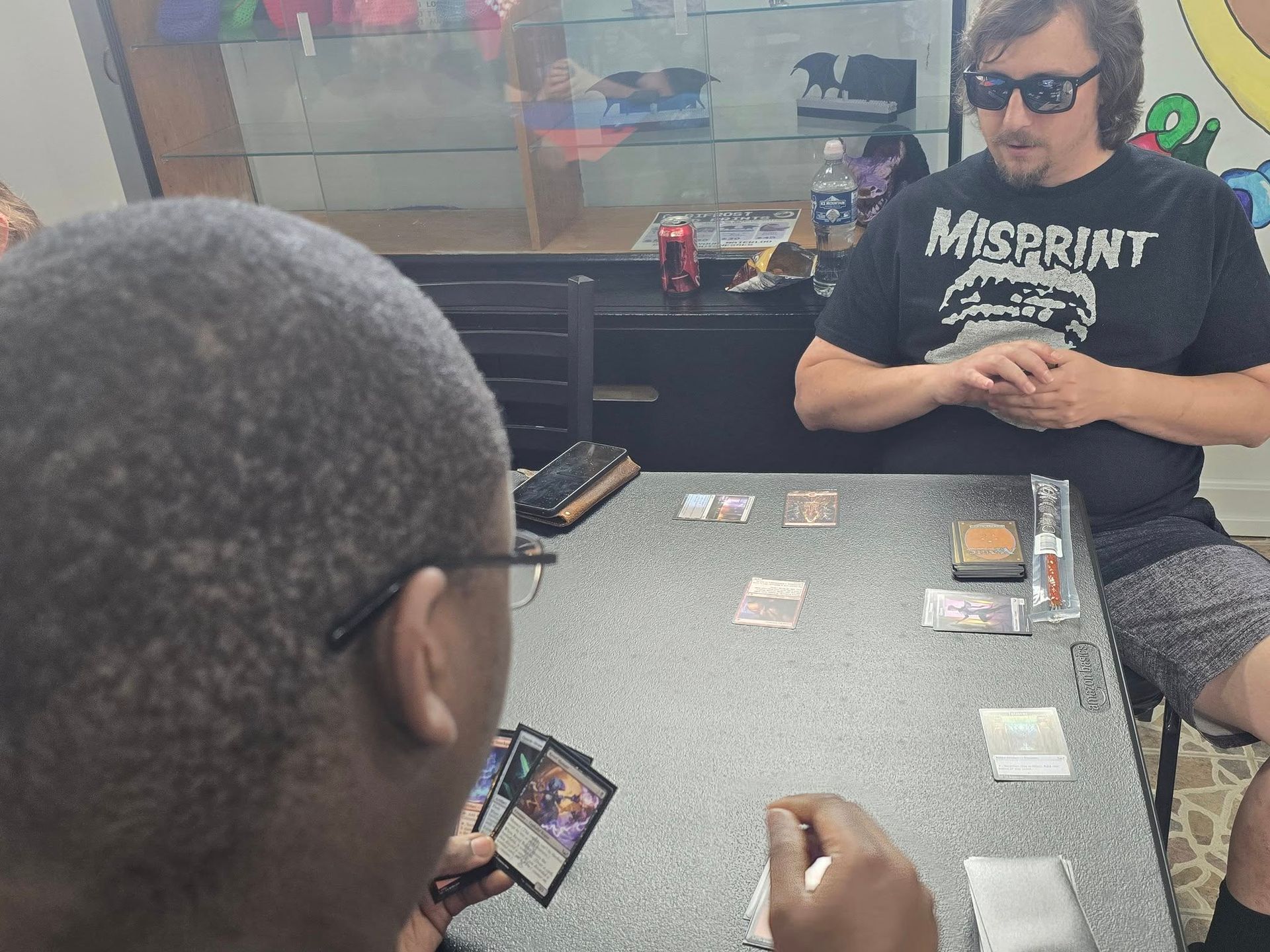 Two men playing Magic: The Gathering. One with glasses, holding cards, the other wearing sunglasses. Cards on a table.