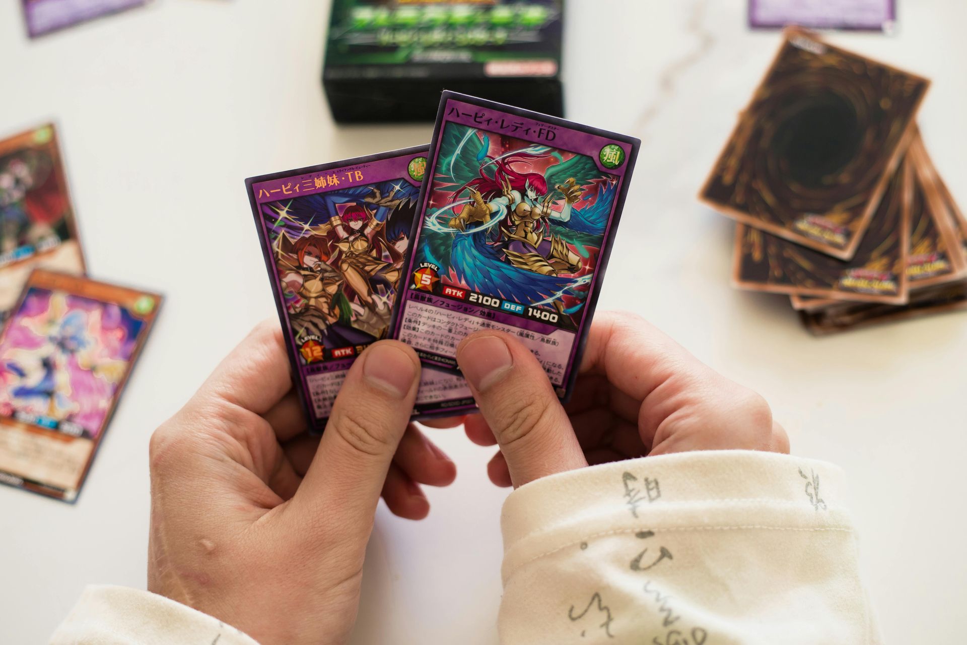 Hands holding two colorful Yu-Gi-Oh! cards, a deck box and cards on a white surface.