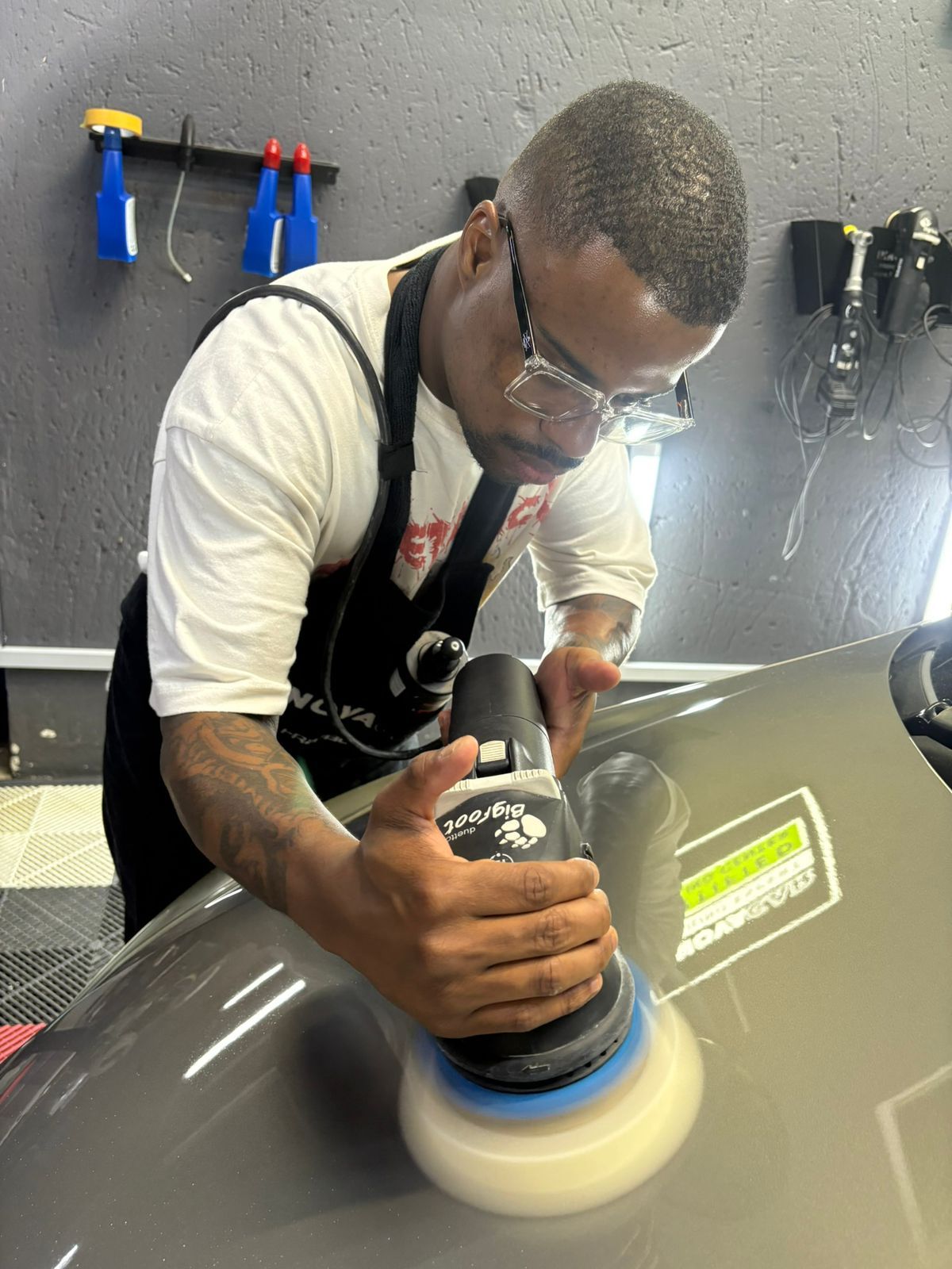 A person polishes a car with a machine in a workshop, wearing safety glasses and an apron.